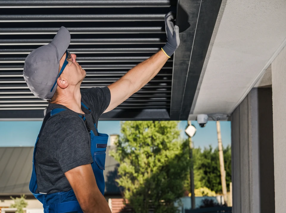 Ouvrier en salopette rouge et casquette installant ou réparant un store à lamelles métalliques sur une terrasse couverte
